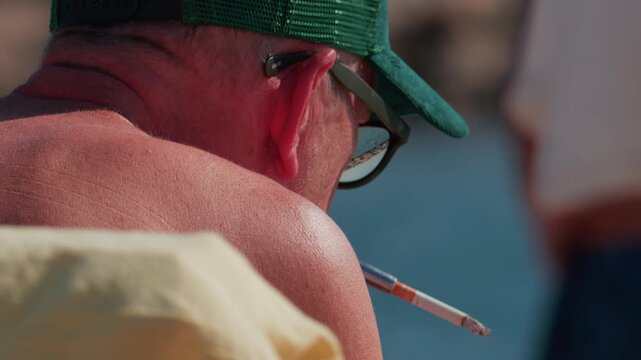 Cannes, France - October 10, 2025: Close up of an elderly man wearing a green cap and sunglasses, smoking a cigarette while sitting by the sea