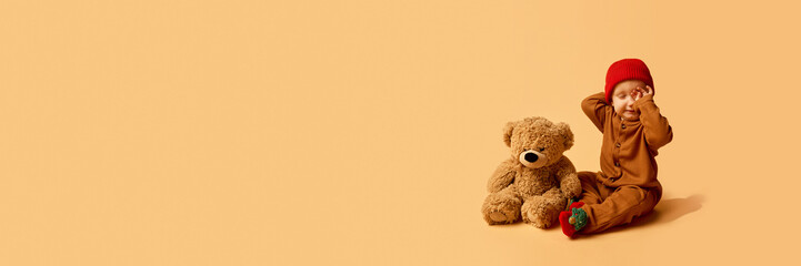 Baby in red hat sitting with teddy bear yawning sleepily on warm monochrome background. Concept of...