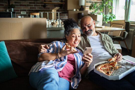 Middle aged couple eating pizza and using a phone after moving in their new home