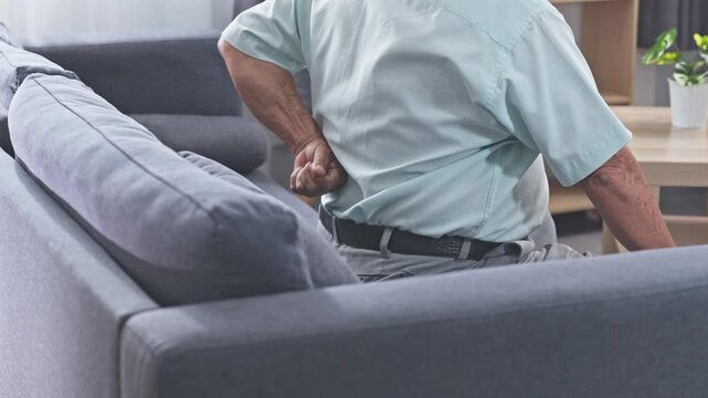 Asian elderly man with lower back pain sitting on sofa holding lumbar in living room showing discomfort posture strain senior cane rehabilitation home scene with soft natural light