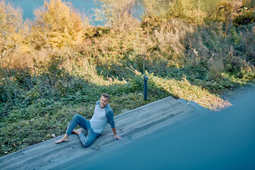 Fototapeta premium Man smiling, relaxing barefoot in pajamas on outdoor wooden stairs, enjoying morning peace and nature