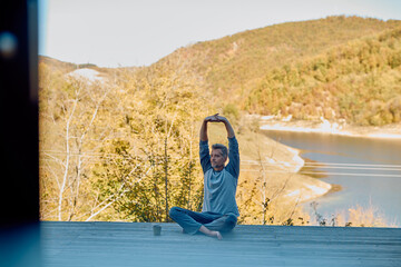 Man with gray hair stretching his arms up, sitting cross-legged on a wooden deck by a lake, surrounded by nature to find peace