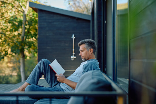 Mature man reading on a modern patio sofa, enjoying peaceful solo time outdoors in sunlight and greenery, relaxed and thoughtful