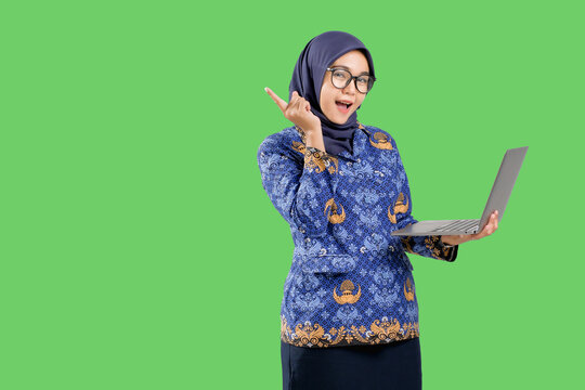 Beautiful Indonesian teacher in blue batik korpri civil servant uniform holding and pointing with laptop against green background, concept of digital learning and innovation.