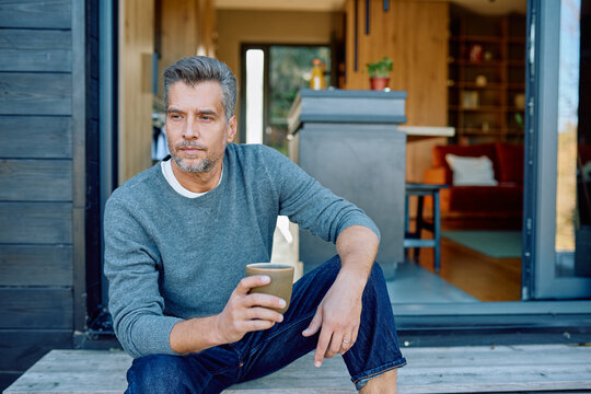 Pensive mature man sitting on porch steps of modern home, holding a cup of coffee and looking away in deep thought