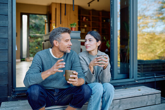 Couple talking and enjoying coffee together on steps outside modern home, feeling happy and connected during leisure time