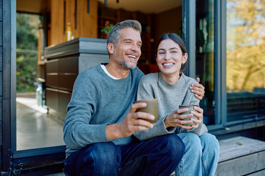 Cheerful couple relaxes, sitting on a wooden porch outside their house, sharing a joyful moment with hot drinks