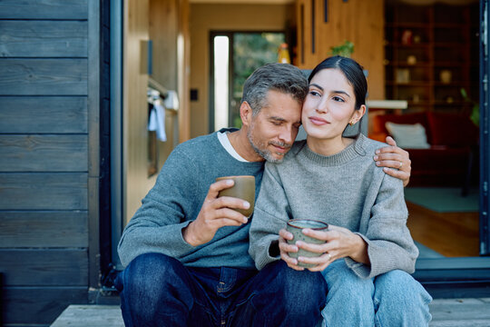 Loving couple sitting on a porch, embracing and holding coffee mugs, finding comfort and connection in a peaceful moment - Powered by Adobe
