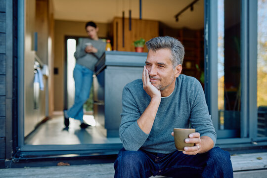 Thoughtful man sitting on porch steps with coffee, smiling, while a woman stands blurred in the background, enjoying home life