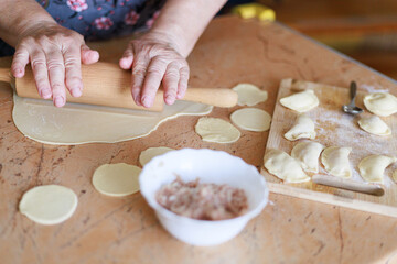Hands of elderly woman rolling dough and shaping dumplings with meat filling on wooden table. Concept of authentic cooking, family tradition, and cozy kitchen life with copy space.