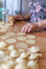 Elderly woman forming dumplings by hand on wooden table surrounded by dough circles. Concept of home cooking, tradition, and warm authentic atmosphere with copy space.