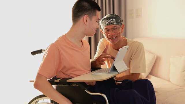 A young man with a disability in wheelchair use tablet for learning with parent in living room, He had tremors similar of Parkinson's disease, Medically-named disability "CP"(cerebral palsy).