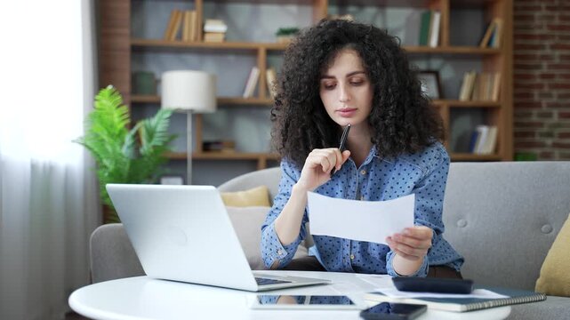 Busy young woman dealing with household finances using website or app on laptop sitting in home office. Conceived adult female holds utility bill and forms payments online by entering data on computer