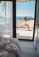 Middle-aged woman doing morning yoga Warrior Pose balance exercise on sea view terace hotel room, doing Virabhadrasana Pose which making your body more balanced and stable. Healthy morning routine.