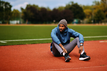 Exhausted African American male athlete sits down on a red running track, taking a determined rest and looking focused after an intense outdoor workout