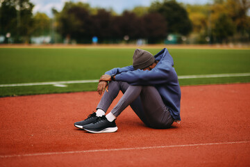 Exhausted  male athlete sits hunched over with his head on his knees on a running track, showing intense fatigue or post-workout recovery.