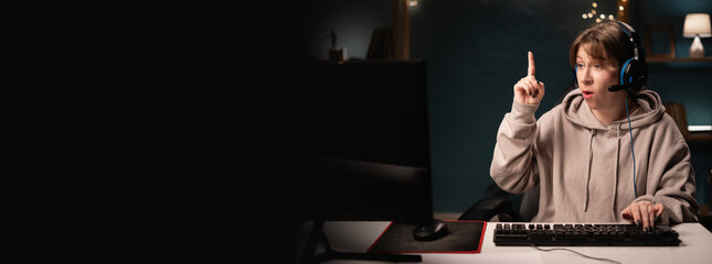 E-sport female gamer in headphones play online video game holding index finger up with great new idea while using computer. Young gaming player broadcast live streaming plays video games