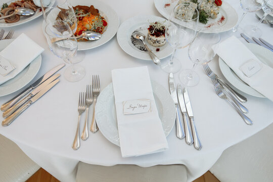 Close-up of an elegant wedding table setting showing a white cloth napkin with a handwritten name card, silver cutlery, a white porcelain plate, and prepared appetizers