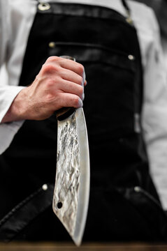A chef or butcher holds a hand-forged knife with a rustic, hammered blade and a wooden handle. The person wears a white chef's coat and black apron. Close-up, selective focus