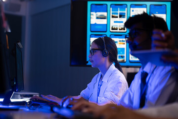 Diverse team of professionals customer service agent wearing headsets and monitoring computer working in dark control room. Female support and assistance call center at night in modern office.