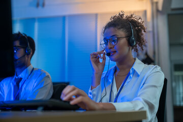 Diverse team of professionals customer service agent wearing headsets and monitoring computer working in dark control room. Female support and assistance call center at night in modern office.