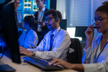 Man support and assistance call center at night in modern office. Diverse team of professionals customer service agent wearing headsets and monitoring computer working in dark control room.