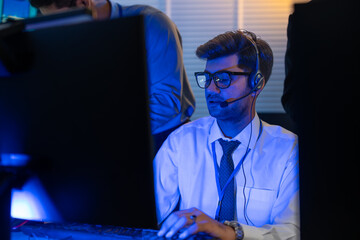 Man support and assistance call center at night in modern office. Diverse team of professionals customer service agent wearing headsets and monitoring computer working in dark control room.