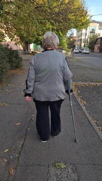 An elderly disabled woman walks with a cane down the street, rear view