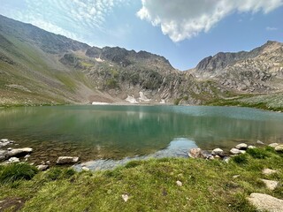 High-altitude mountain lake with clear, turquoise water, surrounded by rocky, snow-dusted slopes. Grassy shore in the foreground. Natural beauty, ecology, hiking