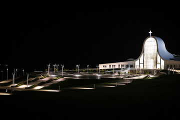 Wide night view of modern illuminated church with glowing cross and surrounding architecture creating spiritual atmosphere and depth.