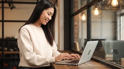Young asian woman smiling while working on a laptop in a cafe by the window - Powered by Adobe