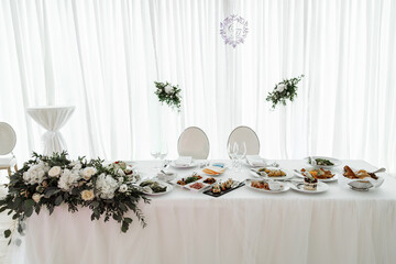 Beautifully decorated wedding reception head table with white roses, lush green floral arrangements, and assorted gourmet appetizers for the bride and groom