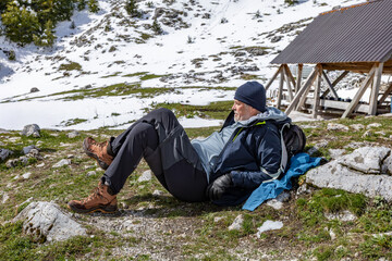 A man resting on a mountain slope with snow patches, dressed in outdoor clothing and hiking boots. He leans back against his backpack, taking a break during a hike in a cold, alpine landscape.