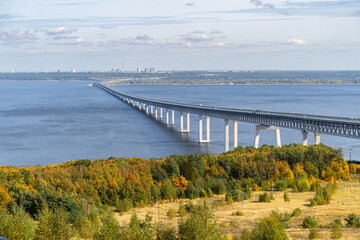 Presidential Bridge in Ulyanovsk spans the Volga River under a clear sky. Autumn foliage and the city skyline create contrast between nature and infrastructure.