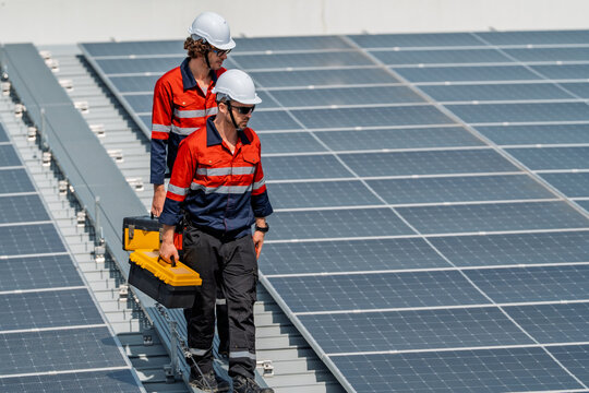 Solar engineer teamwork maintenance energy on rooftop solar panel array under bright sun, two worker in safety helmet and uniform carry toolbox with focus and care during inspection and repair