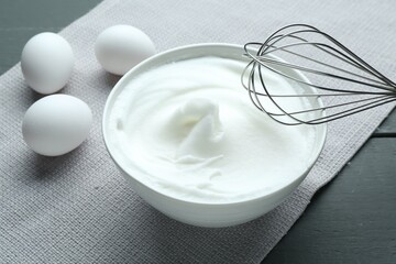 Whipped whites in bowl, whisk and eggs on grey wooden table, closeup