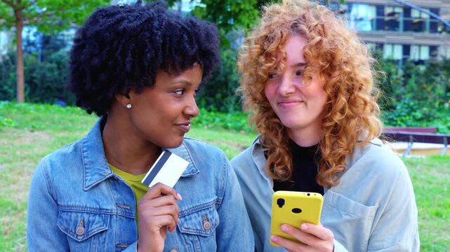  Cheerful African American and Caucasian friends stand in a city park, holding colorful smartphones and a credit card, celebrating cashless convenience, shared shopping success, and digital lifestyle