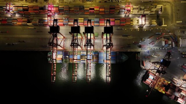 Nighttime aerial view of a busy container terminal with gantry cranes loading a cargo ship at the pier, lights reflecting on calm harbor waters and shipping lanes below