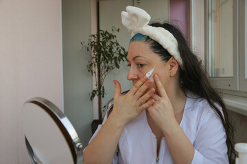 An adult mature woman takes care of her facial skin while sitting in front of a mirror.