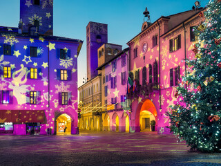Evening festive town square illuminated for the holiday season, historic buildings and medieval towers with light projections and large decorated Christmas tree in Alba, Italy.