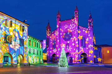 A lit Christmas tree on a cobblestone town square surrounded by illuminated building, in Alba, Italy.