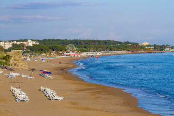 Panoramic view of Side's sandy beach with turquoise water, sunbeds, and sunbathers, backed by the Sorgun forest and resort hotels under a clear evening sky. Antalya, Turkey.

