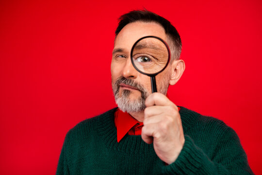 Funny senior man with green sweater peers through magnifying glass in a bright red christmas studio creating a playful festive vibe