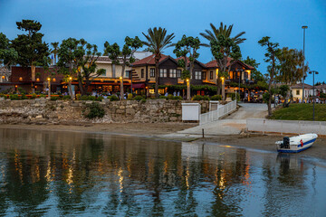Naklejka premium A serene post-sunset view of Side's old town from the water, with illuminated restaurants reflecting in the calm sea and traditional stone houses under a deep blue twilight sky. Antalya, Turkey.