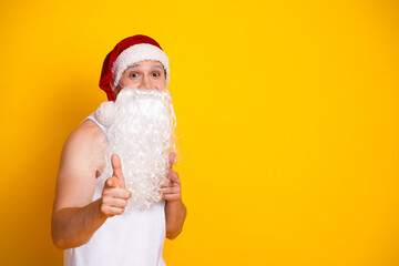 Playful Santa impersonator in white tank top with beard and red hat poses against bold yellow backdrop for festive holiday campaign