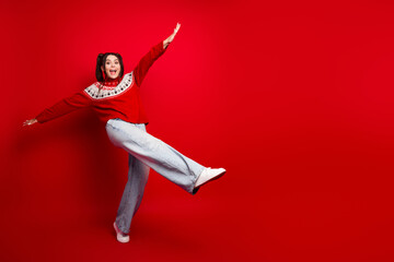 Joyful woman wearing a red christmas sweater kicks with arms outstretched against a bold red background in a festive fashion stock photo