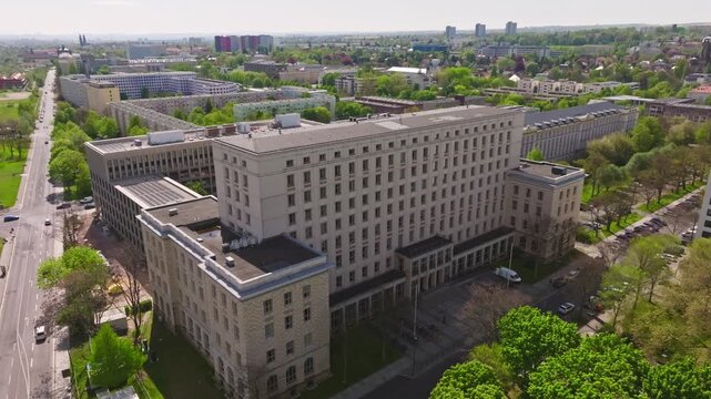 Drone flight over the HTW Dresden on a sunny day. The camera slowly flies backward, revealing greenery, surrounding buildings, cars, and a street from above.
