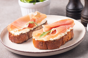Two bruschettas with prosciutto, ricotta cheese and rucola on ciabatta bread. Olive and spices  decorated.