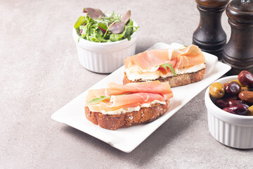 Two bruschettas with prosciutto, ricotta cheese and rucola on ciabatta bread. Olive and spices  decorated.