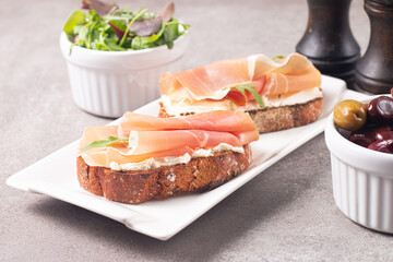 Two bruschettas with prosciutto, ricotta cheese and rucola on ciabatta bread. Olive and spices  decorated.